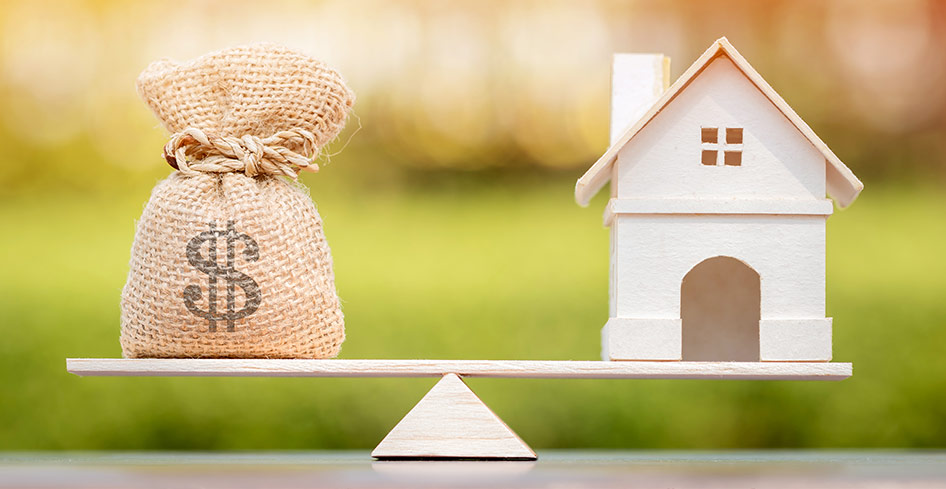 Stock photo of a bag of money and a house sitting on opposite sides of a scale, but they're just made of wood and burlap and natural materials.