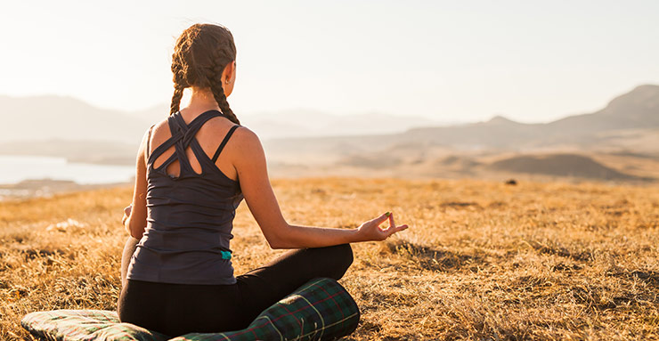 A white woman is doing yoga, sitting cross legged outdoors, facing away from the camera. 