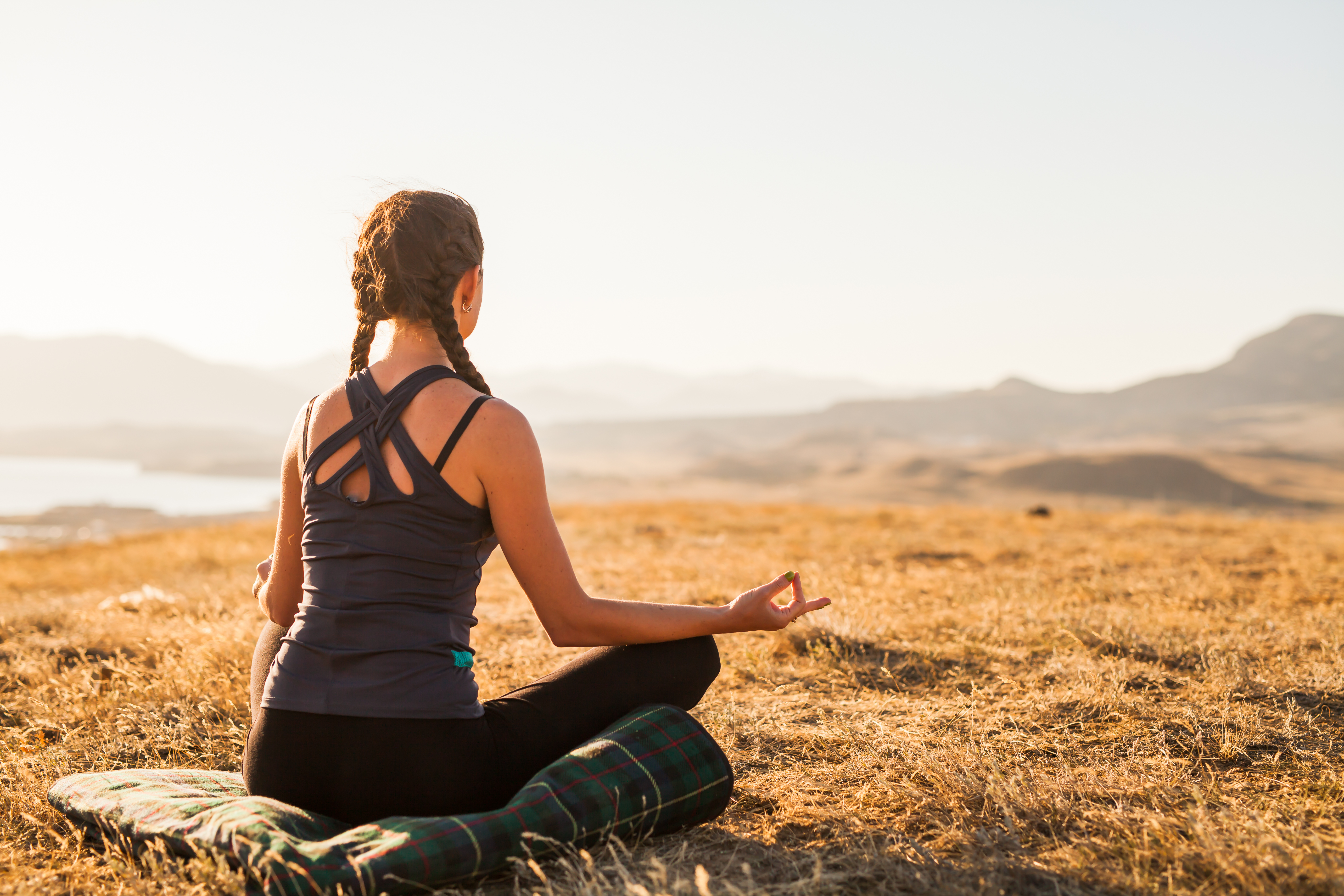 A woman is doing yoga, sitting cross legged outdoors, facing away from the camera. 