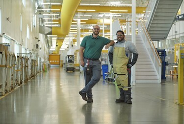 Two men standing in the middle of a bright, well lit manufacturing facility, wearing work gear and smiling.