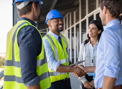 Four people standing together, two of them shaking hands while the other two look on. Two are wearing construction hats and vests and the other two are wearing business attire. Everyone is smiling.