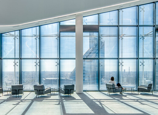 Interior common space of office building with lounge chairs and full smart glass windows from left to right. 
