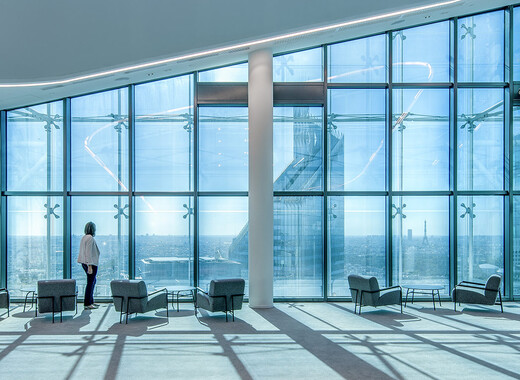 Interior common space of office building with lounge chairs and full smart glass windows from left to right with woman peering out the window.