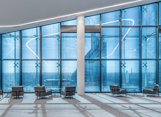 Interior common space of office building with lounge chairs and full smart glass windows from left to right. 
