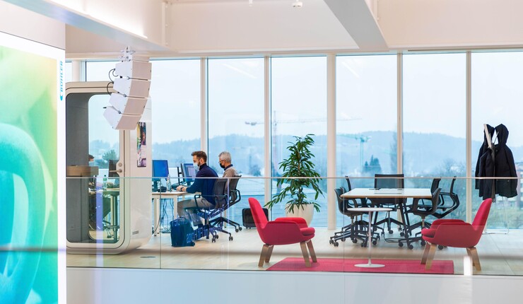 View of inside a modern office, looking at two men working at desks, an open seating area and a table with a stunning set of floor to ceiling windows looking outside with lots of light.