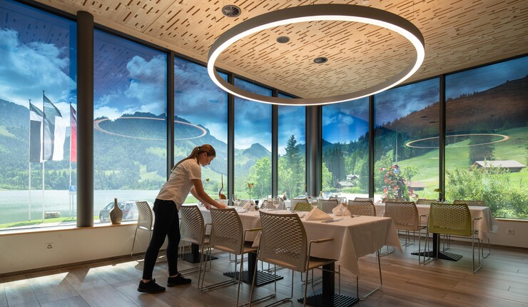 A beautiful interior corner view of a restaurant with a server setting up empty tables. There is glass on all of the exterior walls, showing tinted views of the outdoors.