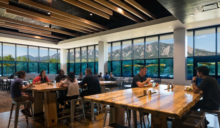 People sitting in an indoor room around tables all at bar stool height. Glass windows all around with beautiful views of nature.